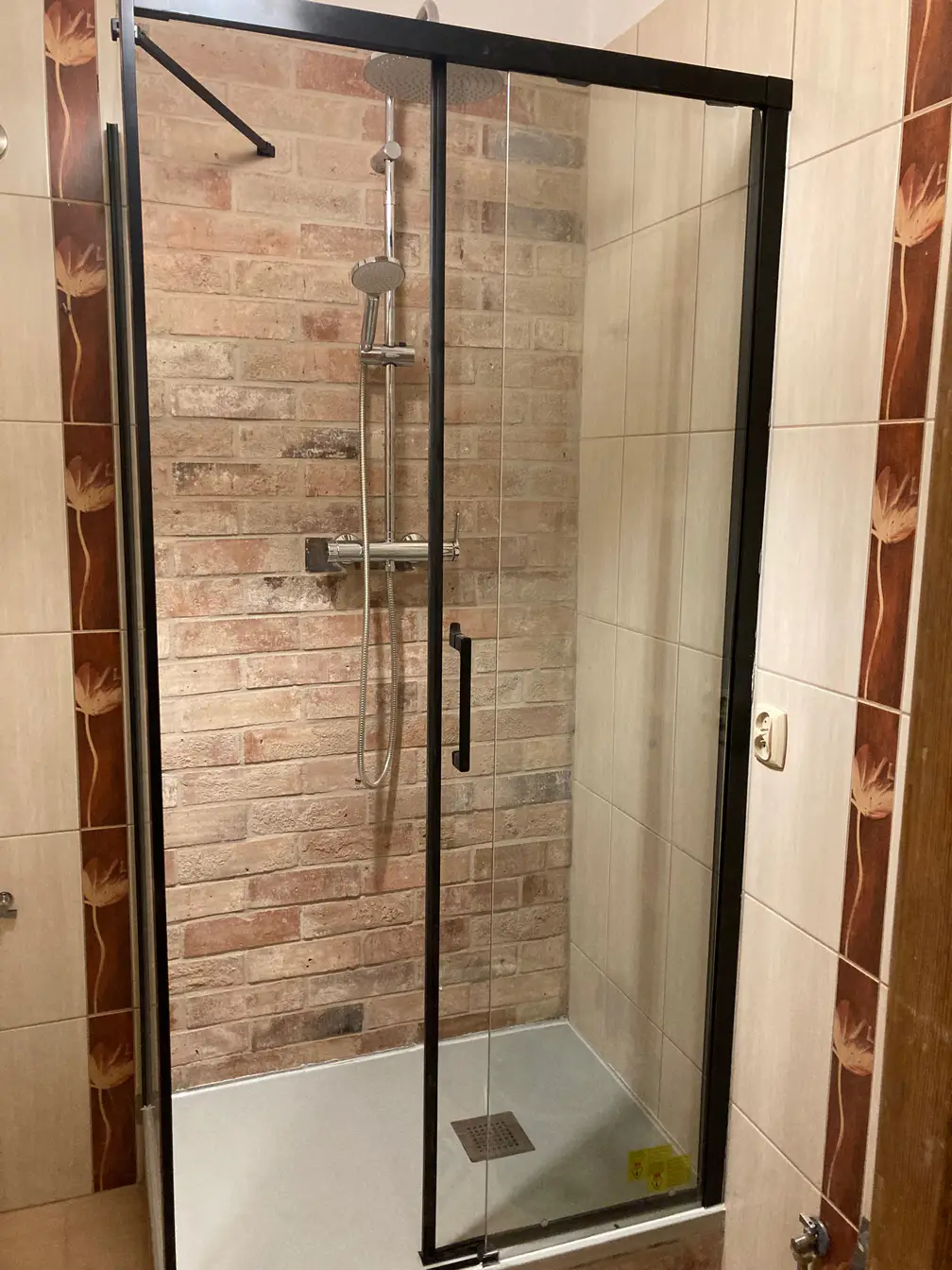 Nowoczesna kabina prysznicowa typu walk-in, gdzie tylna ściana została wykończona zaimpregnowaną starą cegłą, kontrastująca z czarną armaturą.